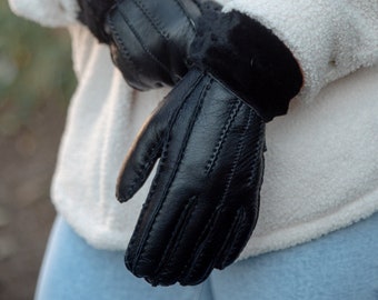 May include: Black leather gloves with a soft, black fur cuff. The gloves have detailed stitching and are worn over a white jacket and blue jeans. The gloves are a winter accessory.