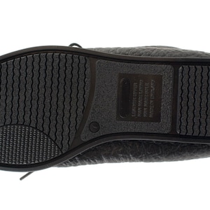 Op de afbeelding: Zwarte leren platte schoenen met een textuurzool en een label aan de zijkant met de tekst "California  Made in China  Leather Upper  Man Made Sole".