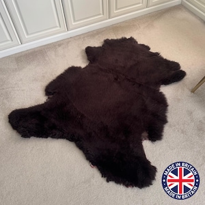 Könnte beinhalten: Ein dunkelbrauner Schaffellteppich mit weicher, strukturierter Oberfläche. Der Teppich ist unregelmäßig geformt und liegt auf einem hellfarbigen Teppich. Das Bild enthält ein "Made in Britain"-Emblem.