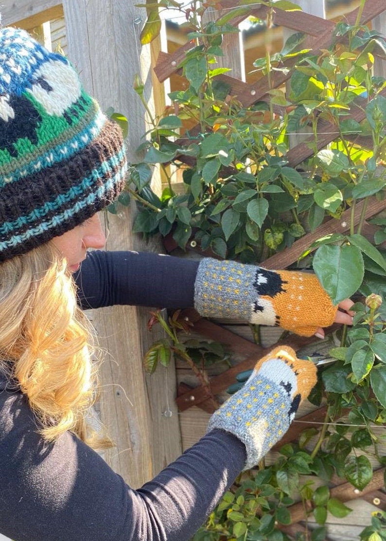 May include: A person wearing a black sweater and a colorful knitted hat with a green, blue, and white pattern is wearing a pair of knitted fingerless gloves with a fox design. The gloves are yellow, orange, gray, and white. The person is pruning a rose bush.