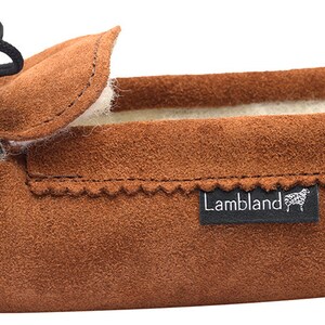 May include: Brown suede moccasin slippers with a black leather tie and a white sheepskin lining. The slippers have a  "Lambland" logo on the side.