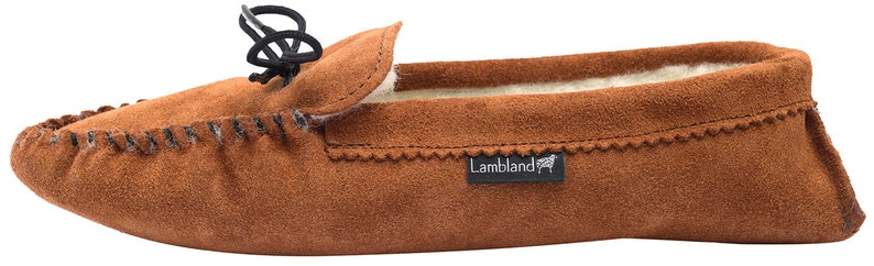 May include: Brown suede moccasin slippers with a black leather tie and a white sheepskin lining. The slippers have a  "Lambland" logo on the side.