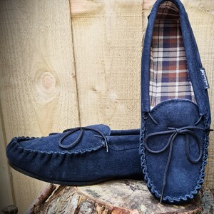 May include: A pair of navy blue suede moccasins with a black ribbon tie. The moccasins have a plaid lining and are sitting on a tree stump.