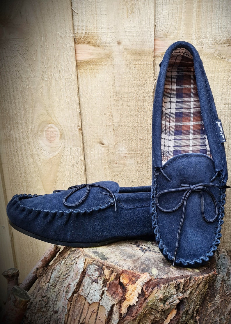 May include: A pair of navy blue suede moccasins with a black ribbon tie. The moccasins have a plaid lining and are sitting on a tree stump.