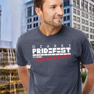Puede incluir: Una camiseta azul marino con el texto "OZARKS PRIDE FEST" en letras blancas. Debajo del texto, la camiseta dice "IT'S GIVING: REVOLUTION" en rojo. La camiseta la lleva una persona.
