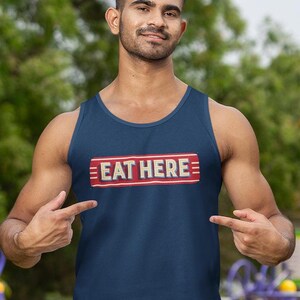 May include: Navy blue tank top with the text "EAT HERE" in a red and gold rectangular design. The tank top is a casual style, suitable for warm weather.