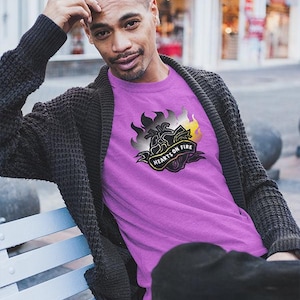 Könnte beinhalten: Eine Person trägt ein lila T-Shirt mit dem Text "HEARTS ON FIRE" in einem Banner-Design. Das Shirt hat eine Grafik von Flammen in Schwarz, Grau und Gelb. Die Person trägt auch eine dunkelgraue Strickjacke und eine schwarze Hose.