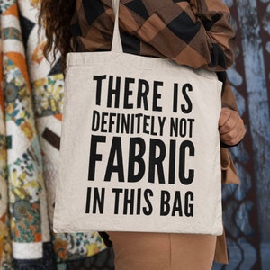 Puede incluir: Bolso tote de color crema con el texto "THERE IS DEFINITELY NOT FABRIC IN THIS BAG" en letras negras en negrita. El bolso es sostenido por una persona que lleva una camisa a cuadros marrón y negro.