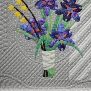 May include: A quilt block featuring a bouquet of purple irises and yellow forsythia flowers. The block is titled "16th and Baltimore, Block 1: Iris and Forsythia" and is 18 inches by 18 inches.