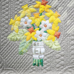 May include: A quilt block featuring a bouquet of yellow daffodils and white flowers in a green vase. The block is titled "Daffodil and Lambs Ear" and is part of the 16th and Baltimore quilt series. The block is 18 inches by 18 inches.