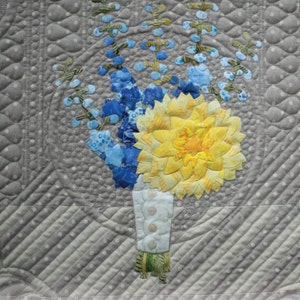 May include: A quilt block featuring a yellow dahlia and blue delphiniums in a white vase. The block is part of the 16th and Baltimore quilt pattern, Block 7: Dahlia and Delphiniums, ECQ 16B07, 18 inches by 18 inches.