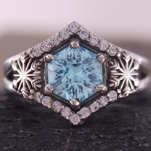 May include: A silver ring with a hexagonal blue gemstone surrounded by smaller clear gemstones. The ring features snowflake designs on either side of the gemstone.