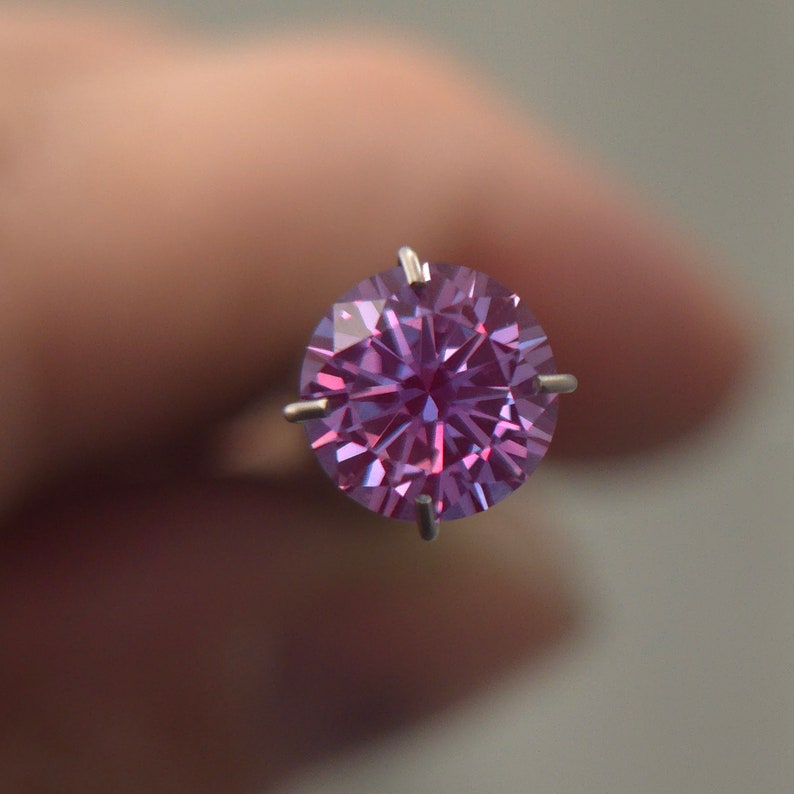 May include: A round, faceted, purple gemstone with a star-like pattern in the center, held in a prong setting.