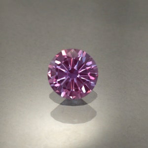May include: A round, faceted gemstone with a purple and pink hue. The gemstone is set on a white background.