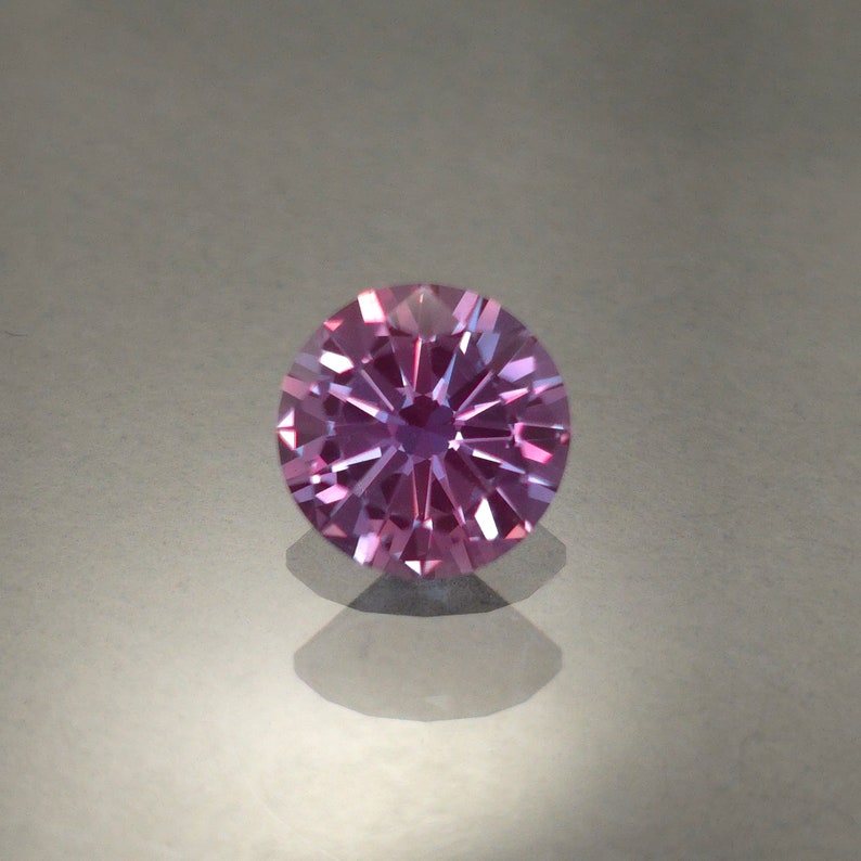 May include: A round, faceted gemstone with a purple and pink hue. The gemstone is set on a white background.