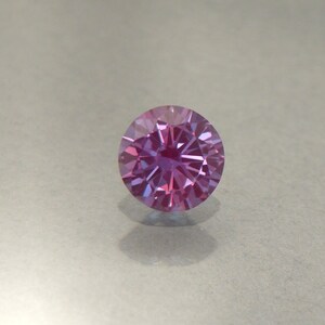 May include: A round, faceted gemstone with a pink and purple hue. The gemstone is set in a clear, round setting.