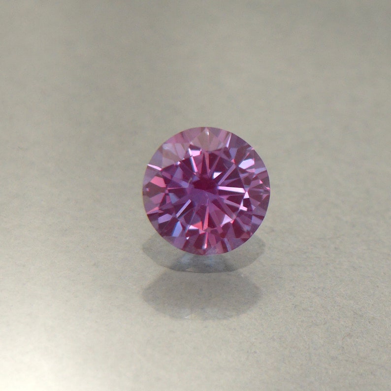 May include: A round, faceted gemstone with a pink and purple hue. The gemstone is set in a clear, round setting.