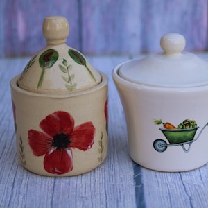 May include: Two ceramic jars with lids. One is beige with a red poppy design and green accents. The other is white with a wheelbarrow illustration. Both jars have a round knob on top.