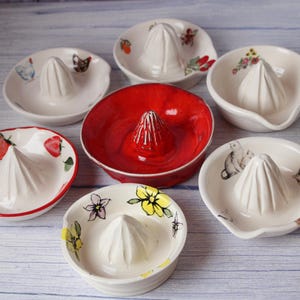 May include: A selection of ceramic citrus juicers. The white juicers have a central reamer and are decorated with hand-painted designs of strawberries, flowers, and butterflies. One juicer is a solid red colour.