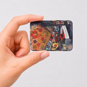 May include: A rectangular magnet featuring a vibrant, multi-colored artistic illustration of a street scene. The artwork includes red floral patterns, various small buildings, and a white street sign clearly displaying the text Mink Mahalo Rd.