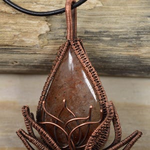 May include: A copper wire wrapped pendant featuring a brown gemstone with a teardrop shape. The pendant is suspended from a black leather cord and has a copper wire lotus design at the bottom.