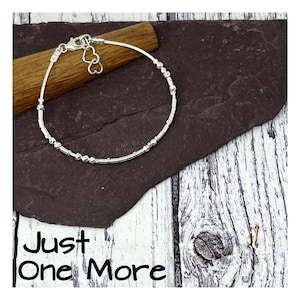 May include: A silver bracelet with small beads and a heart charm. The bracelet is on a wooden surface with the text "Just One More" below it.