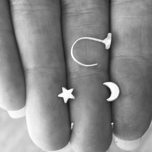 May include: Three silver nose rings in the shape of a crescent moon, a star, and a hoop. The rings are displayed on a hand.