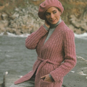 以下が含まれることがあります： ピンクのニットカーディガン。ショールカラー、リボンベルト、ポケット付き。グレーのタートルネックセーターの上に着ています。
