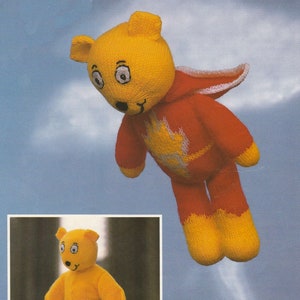 May include: A yellow knitted teddy bear wearing a red knitted cape with white accents. The bear is flying with a trail of white smoke behind it. A smaller image of the same bear without the cape is shown in the bottom left corner.