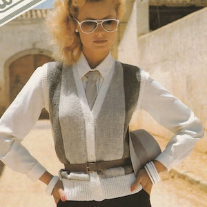 Pode incluir: Uma mulher vestindo uma camisa branca abotoada com um colete de malha listrado cinza e branco. O colete tem um decote em V e um fecho de botão. Ela está usando um cinto de couro marrom e calças pretas. Ela também está usando óculos de sol brancos e um chapéu branco.
