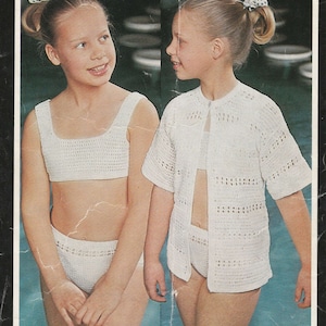 May include: Two girls wearing white crocheted swimwear. The girl on the left is wearing a halter top and matching bikini bottoms. The girl on the right is wearing a cardigan and matching bikini bottoms.