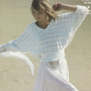 May include: White crocheted sweater with a v-neck and a loose fit. The sweater is shown on a model wearing a white skirt.