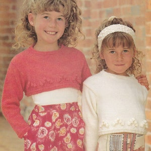 May include: Two young girls wearing knitted sweaters with crocheted trim. The girl on the left is wearing a pink sweater and a floral skirt. The girl on the right is wearing a white sweater and a patterned skirt.