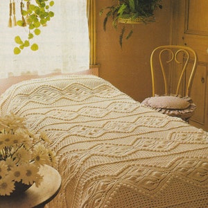 May include: A white crocheted bedspread with a textured pattern of waves and diamonds. The bedspread is on a bed with a white headboard and a white pillow. A chair with a floral cushion is in the background.