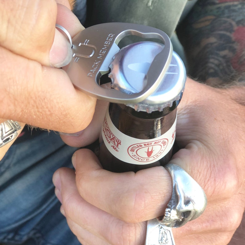 May include: A silver bottle opener with a key ring and the text "Pack Member" on it. The opener is being used to open a brown glass bottle of beer.