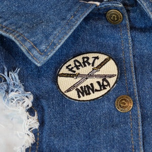 May include: A close-up of a blue denim jacket with a white and black embroidered patch that says "Fart Ninja" with two crossed swords.