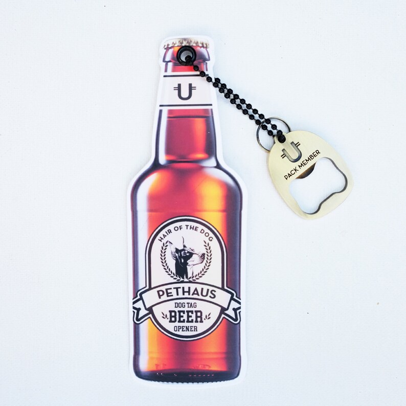 May include: A brown beer bottle shaped dog tag bottle opener with the text "Hair of the Dog" and "Pethaus Dog Tag Beer Opener". The bottle opener is attached to a gold metal bottle opener with the text "U Pack Member".