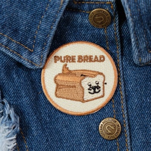 May include: A white and brown embroidered patch with the text "Pure Bread" and a cartoon image of a loaf of bread with a face. The patch is sewn onto a blue denim jacket.