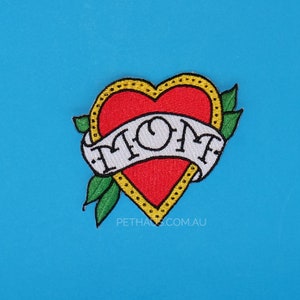 May include: Red heart with a yellow outline and a white banner that says "MOM" in a traditional tattoo style. The heart has green leaves on either side.