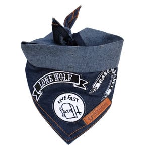 May include: Dark blue denim dog bandana featuring embroidered patches with the phrases "Lone Wolf", "Babe Magnet", and "Live Fast Holes". It has a lighter blue rolled edge and orange stitching. A brown leather tag is attached.