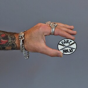 May include: A white round patch with black embroidered text that reads "Fart Ninja" and two crossed swords.