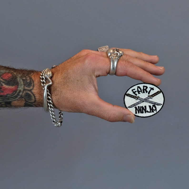 May include: A white round patch with black embroidered text that reads "Fart Ninja" and two crossed swords.