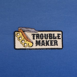 May include: A beige embroidered patch with a black border. The patch features a black and brown switchblade knife with a yellow brass knuckle. The text "Trouble Maker" is embroidered in black on the patch. The text "pethaus.com.au" is embroidered in black at the bottom of the patch.