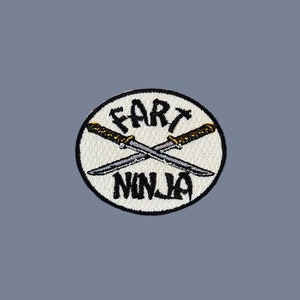 May include: A white circular patch with black embroidered text that reads "Fart Ninja" and two crossed swords with gold handles.