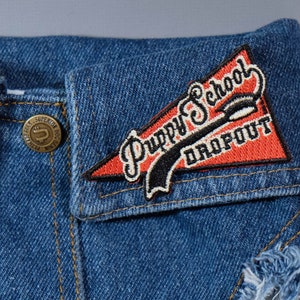 May include: A red, white, and black embroidered patch with the text "Puppy School Dropout" on a blue denim jacket.