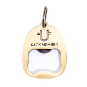 May include: Gold-coloured metal bottle opener keychain with a bone-shaped opening. The keychain has a ring at the top and the words "PACK MEMBER" and "U" engraved on the front.