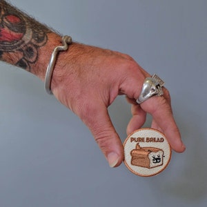 May include: A round, beige embroidered patch with the text "PURE BREAD" and a cartoon image of a loaf of bread with a smiling face.