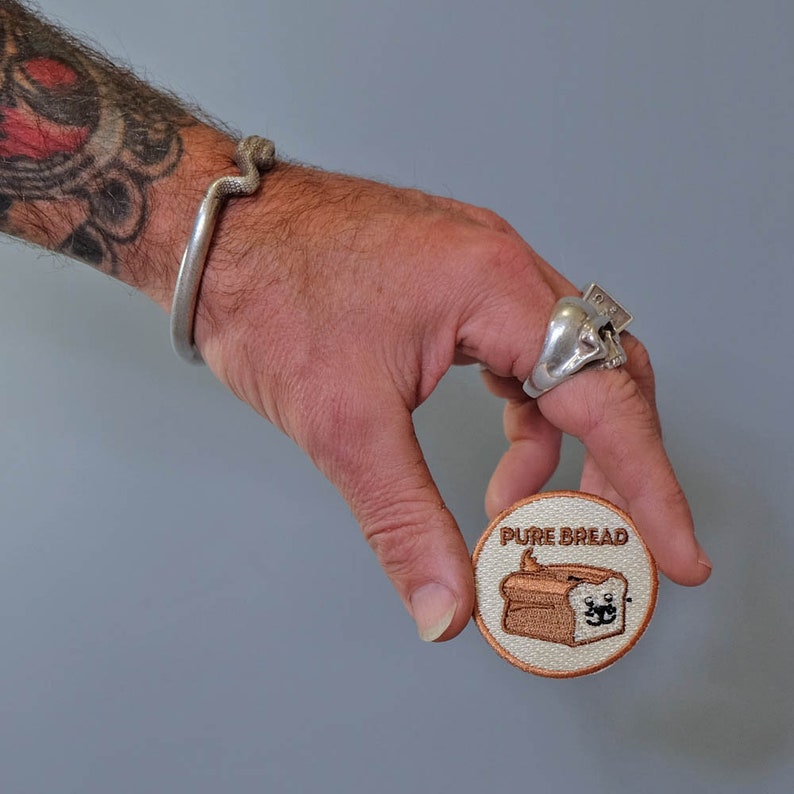 May include: A round, beige embroidered patch with the text "PURE BREAD" and a cartoon image of a loaf of bread with a smiling face.