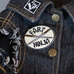 May include: A white, round patch with black embroidery that says "Fart Ninja" with two crossed swords. The patch is sewn onto a dark denim jacket.