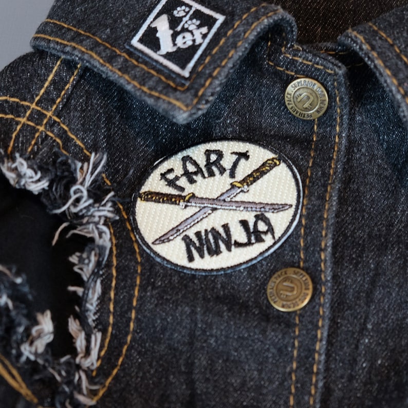 May include: A white, round patch with black embroidery that says "Fart Ninja" with two crossed swords. The patch is sewn onto a dark denim jacket.